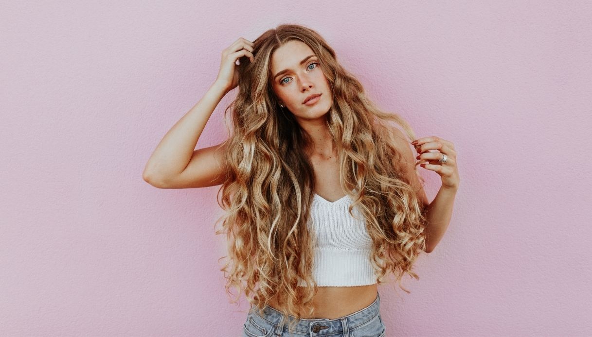 ragazza bionda occhi azzurri con capelli lunghissimi mossi top canottiera bianca e jeans su sfondo rosa