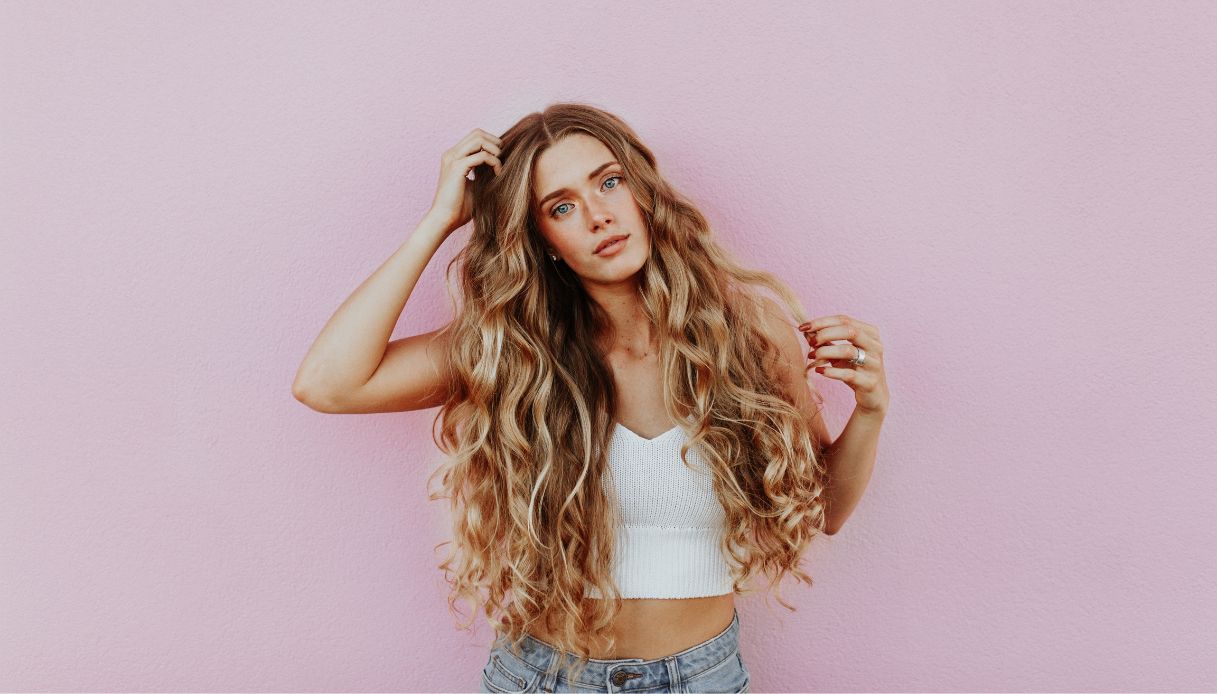 ragazza con capelli biondi mossi su sfondo rosa con jeans e top bianco che si tocca la testa con una mano