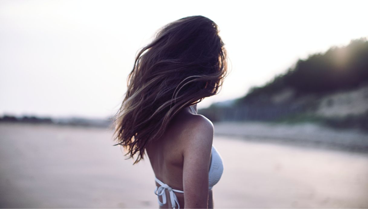 ragazza di spalle con costume bianco e capelli castani al vento con sfondo spiaggia