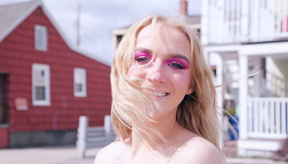 ragazza biondo platino sorridente con capelli sulla faccia trucco rosa fucsia e sfondo casa rossa