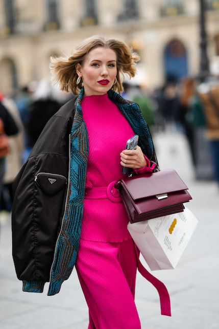 Modella con abito fucsia, cappotto nero, borsa scura e capelli al vento
