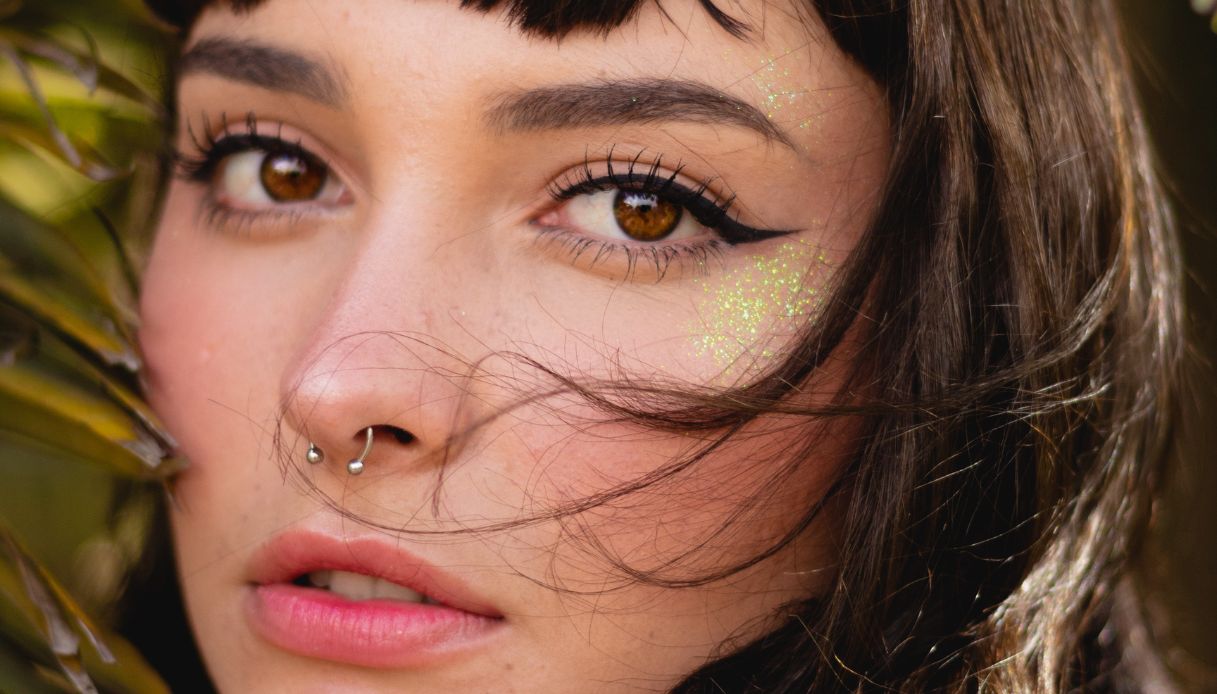 primo piano ragazza mora con frangia, eyeliner, septum al naso, glitter sugli zigomi e rossetto chiaro