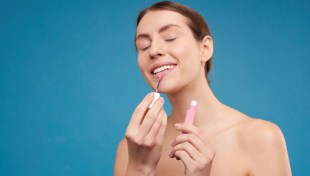 ragazza sorridente mora con capelli raccolti applica pennello sulle labbra per mettere rossetto su sfondo azzurro