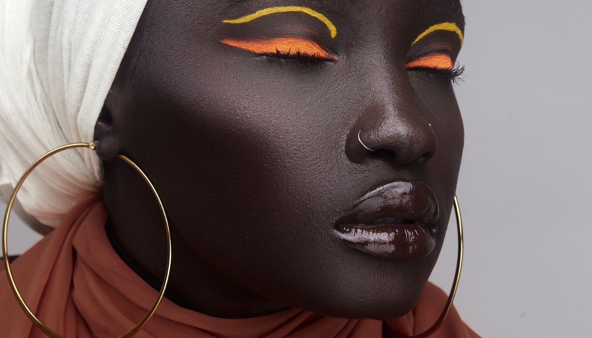 donna pelle molto scura con orecchini grandi cerchi dorati trucco con eyeliner arancione e giallo e labbra con gloss scuro