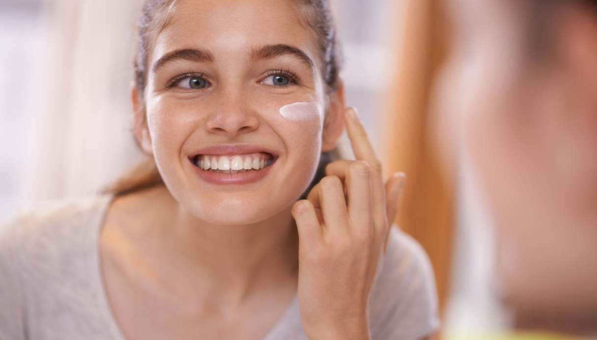 Una ragazza con i capelli legati si guarda allo specchio e sorride mentre applica la crema idratante