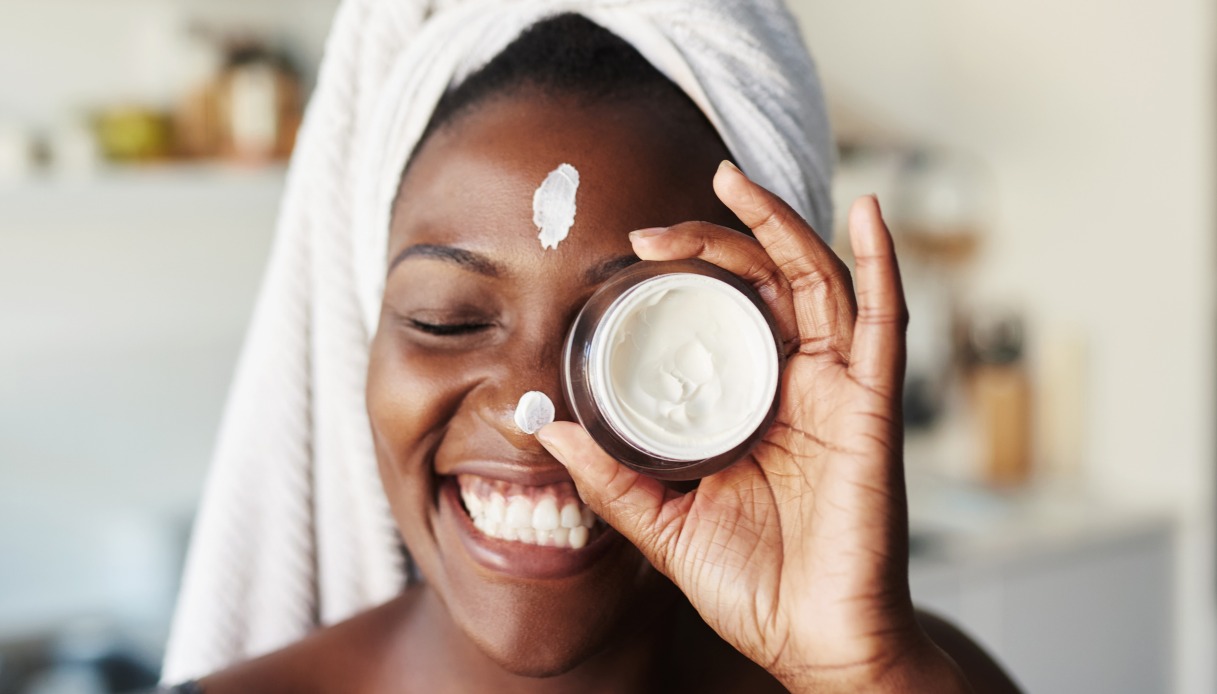 Ragazza nera con turbante bianco sorride a occhi chiusi con della crema sul viso e con il vasetto tenuto tra le mani davanti all'occhio sinistro
